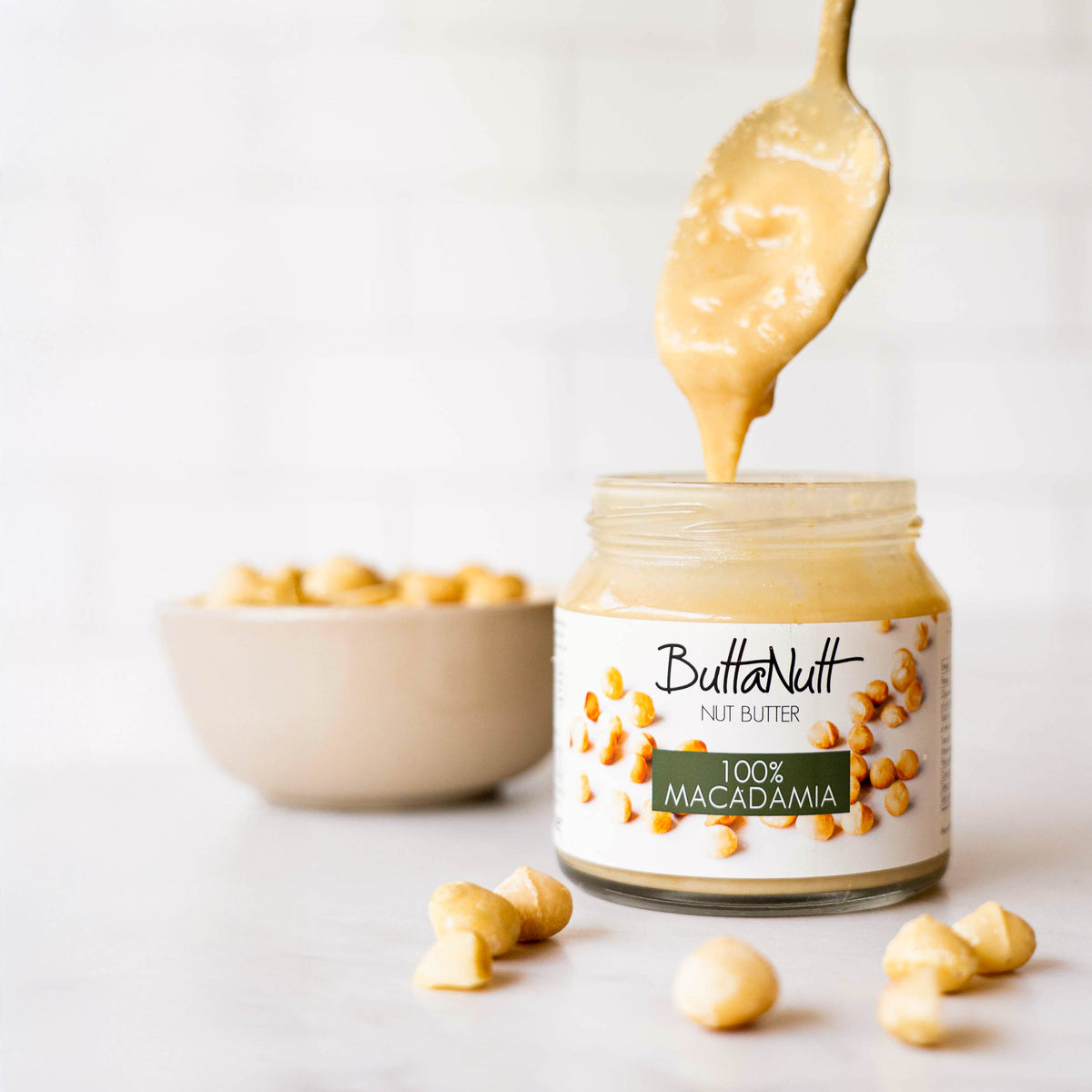 A spoon lifting creamy 100% Macadamia nut butter from a ButtaNutt jar, with whole macadamias beside it