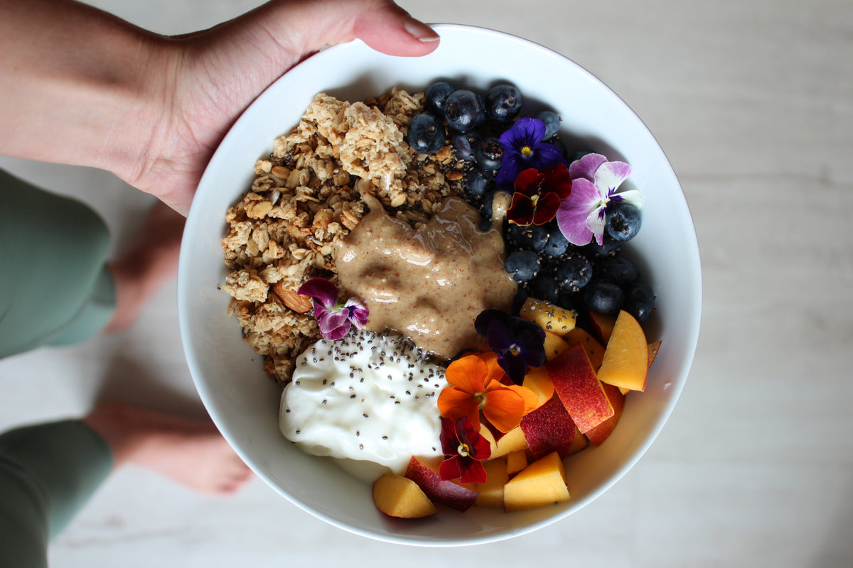 A hand holding a bowl of granola, yogurt, blueberries and peach topped with a spoonful of macadamia nut butter