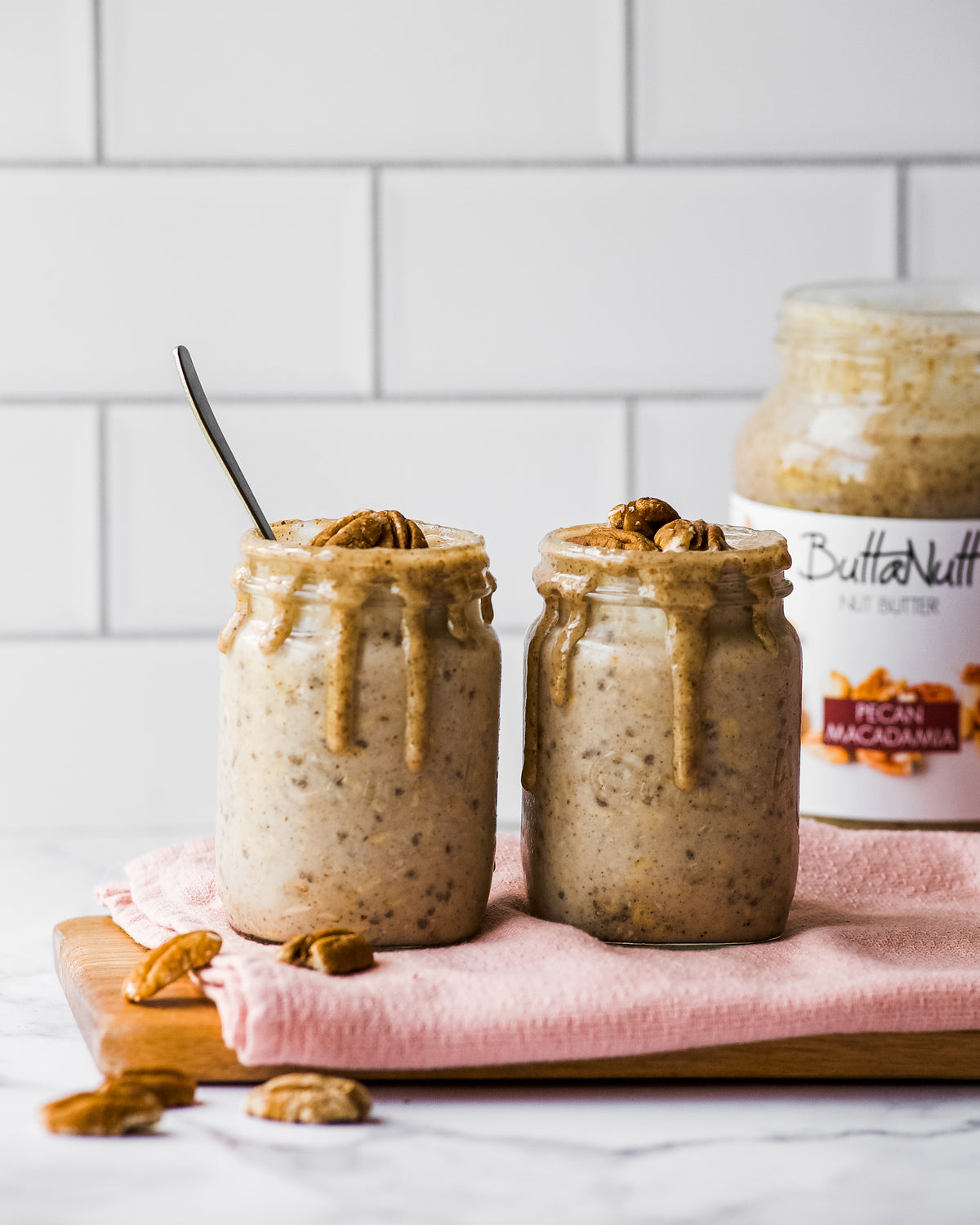 Two jars of Pecan Macadamia nut butter overnight oats topped with pecans, with a jar of ButtaNutt Pecan Macadamia behind